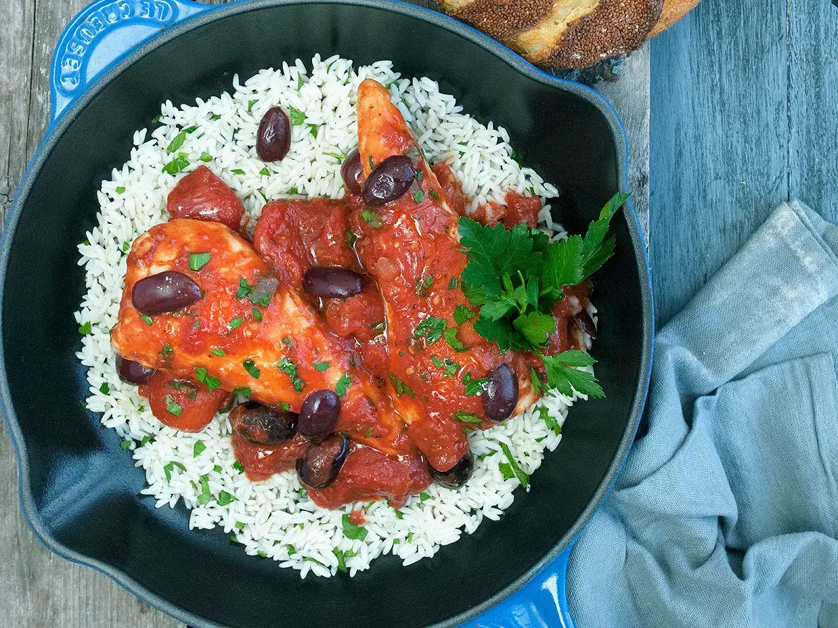 Un plat de poulet sauce tomate savoureux, servi sur un lit de riz blanc, garni d'olives noires et de persil frais.