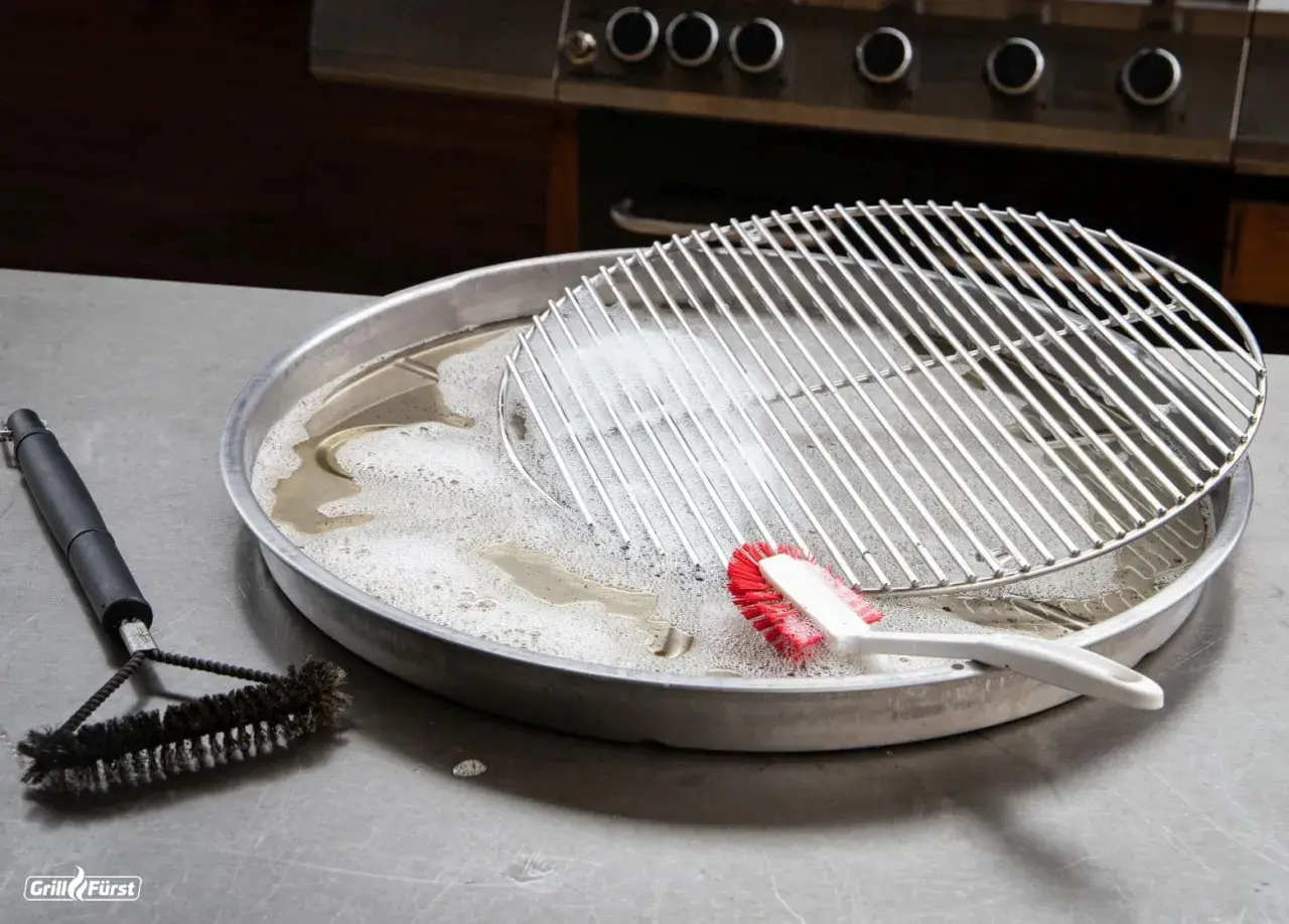 Grillrost mit eingebrannten Resten wird in Seifenlauge eingeweicht. Eine Bürste liegt daneben, bereit, den eingebrannten Grillrost zu reinigen.