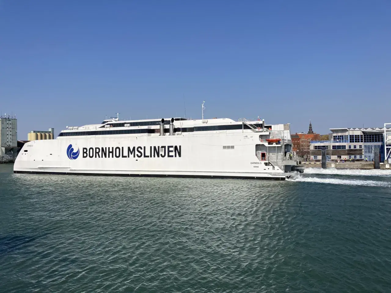 Biały prom "Bornholmslinjen Express" wypływa z portu, gotowy na podróż do Szwecji. W tle widać zabudowania portowe.