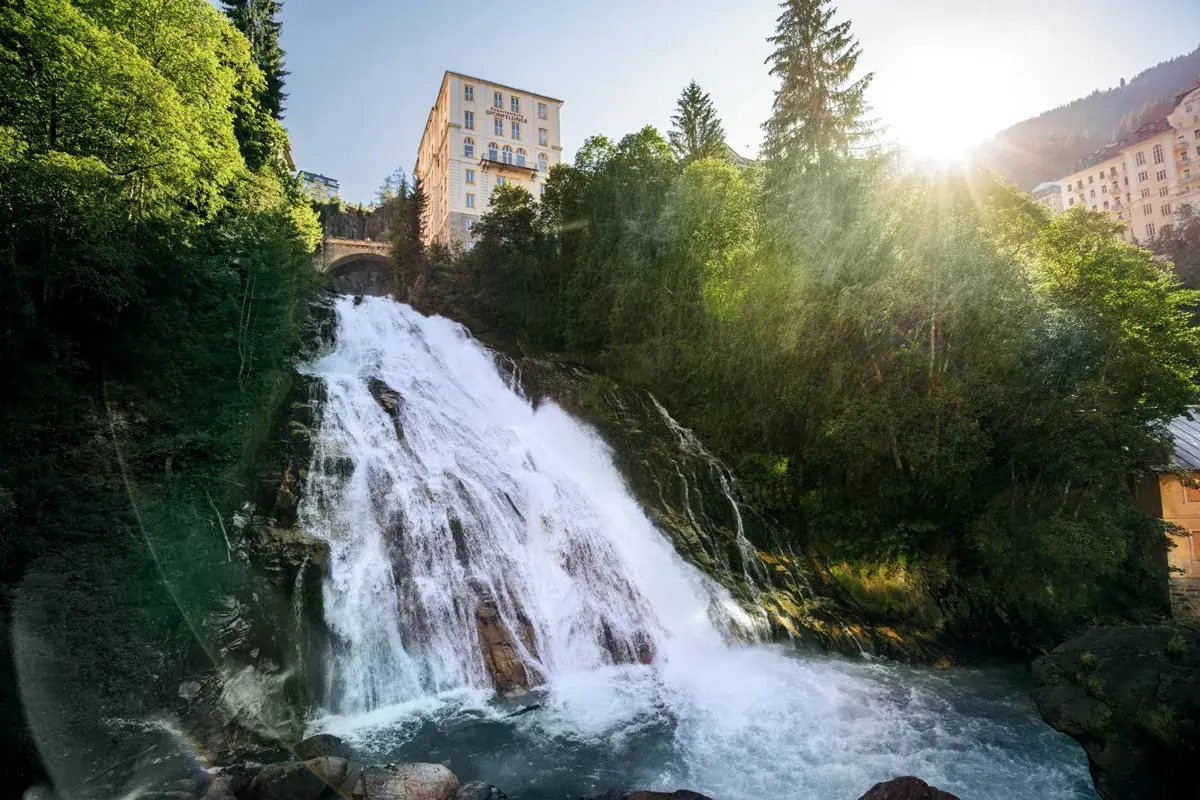 Tosender Wasserfall stürzt durch grüne Hänge in Bad Gastein Altstadt, wo sich ein imposantes Hotel über dem Naturschauspiel erhebt.