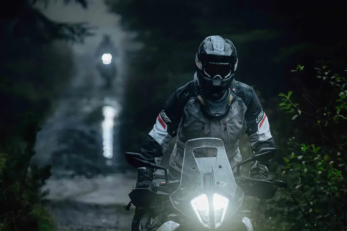 Motocyklista w kasku i kurtce jedzie po mokrej, leśnej drodze w nocy.
