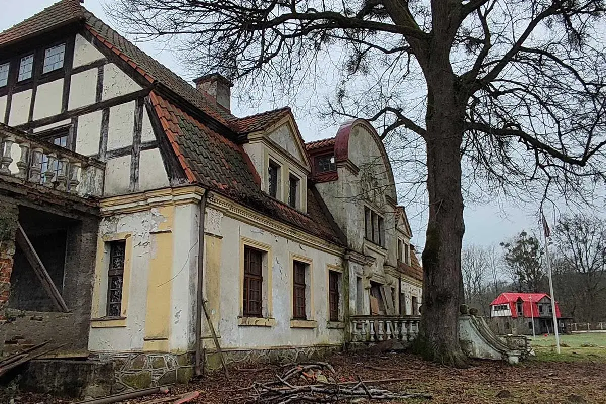 Zniszczony dworek w Paraszyne, z charakterystyczną architekturą i opadającą fasadą, otoczony drzewami.