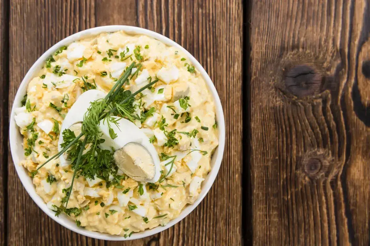 Insalata cremosa con uova di quaglia sode, erbe fresche e un uovo intero a metà come guarnizione, servita in una ciotola bianca su un tavolo di legno rustico.