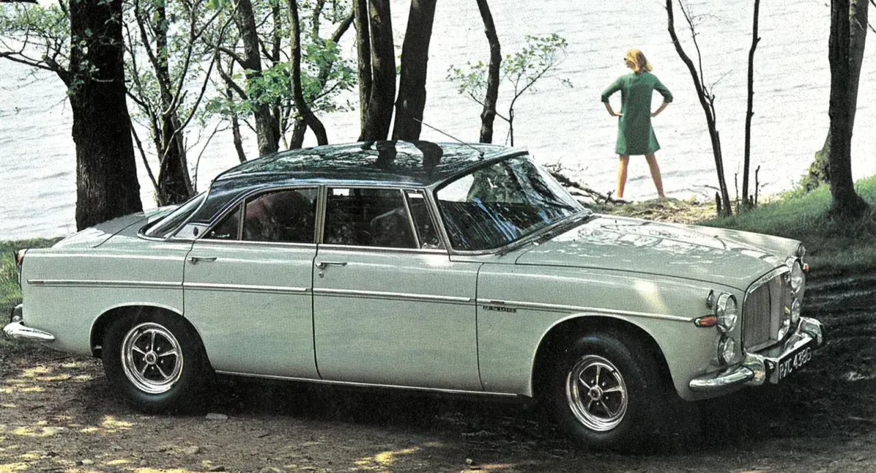 En ljusgrå Rover P5 står parkerad vid en sjö. En kvinna i grön klänning står vid vattenbrynet.
