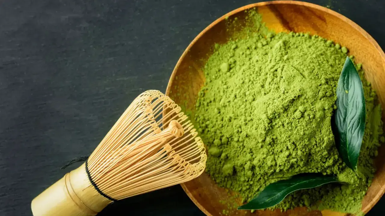 Ein hölzerner Löffel mit feinem, leuchtend grünem Matcha-Pulver und ein Bambusbesen daneben. Der Geschmack ist intensiv, leicht bitter.