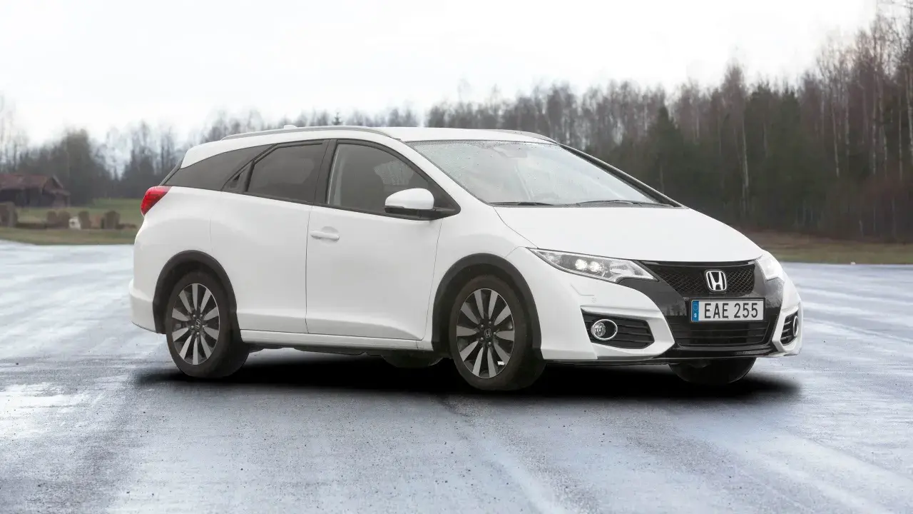 Vit Honda Civic Tourer parkerad på en fuktig väg med träd i bakgrunden.
