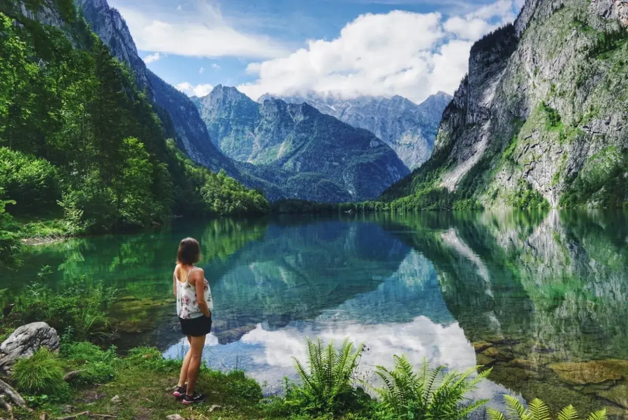 Frau blickt auf spiegelglatten See in den bayerischen Regionen, umgeben von steilen, bewaldeten Bergen.