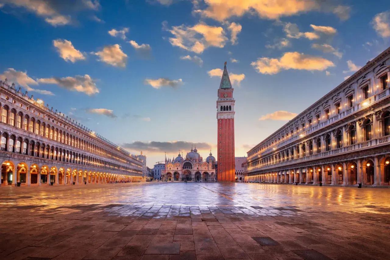 Le **place Saint-Marc à Venise** s'illumine au crépuscule. La basilique et le campanile se dressent majestueusement sous un ciel nuageux.