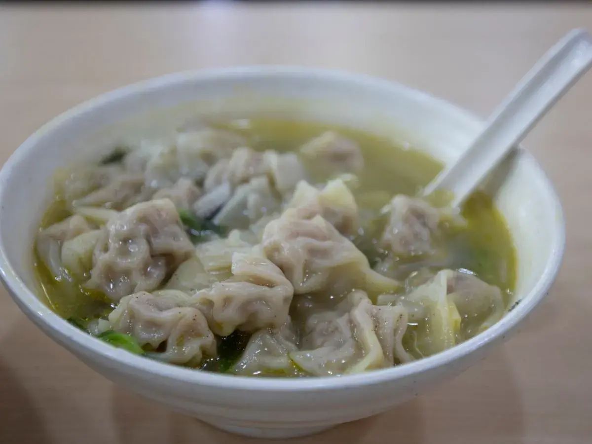 A miseczka pełna aromatycznej zupy wonton, z widocznymi pierożkami i zielonymi warzywami. Łyżka zanurzona w bulionie.