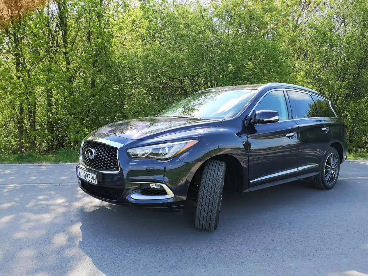 Ciemny Infiniti QX60 zaparkowany na poboczu drogi, otoczony zielenią drzew.