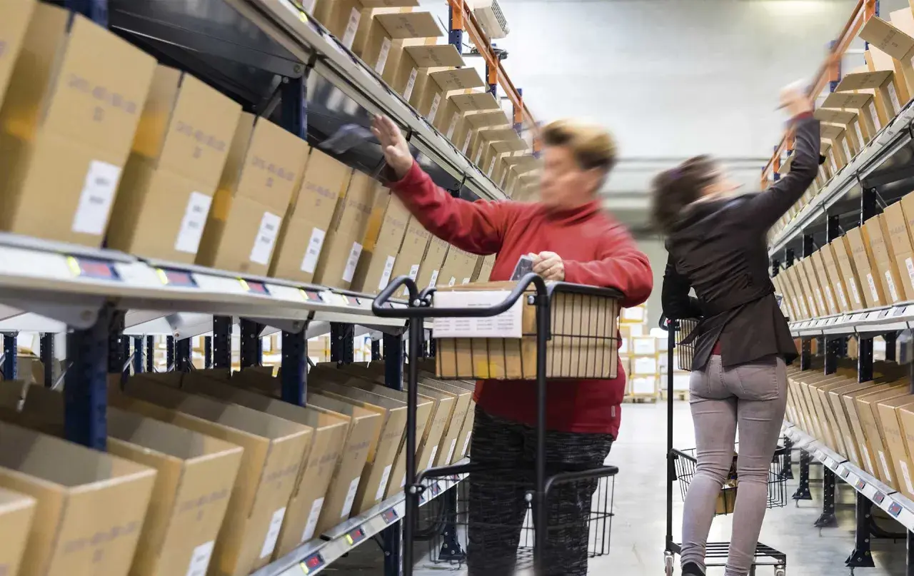 Mitarbeiter im Lager führen pick & pack aus, greifen Kartons aus Regalen und legen sie in Wagen.