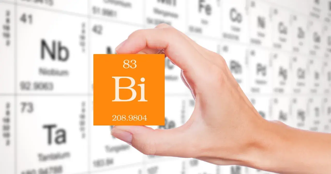 Dłoń trzyma pomarańczową kartkę z symbolem pierwiastka bizmut (Bi) i jego liczbą atomową 83. W tle widać tablicę Mendelejewa.