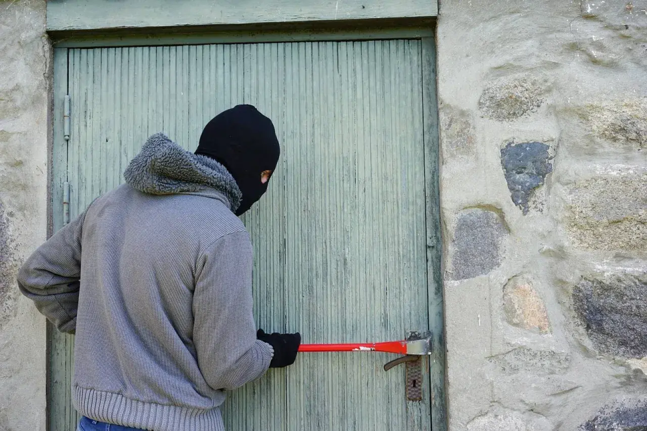 Un individu masqué tente de forcer une porte avec un pied-de-biche, acte relevant du cambriolage et du code pénal.