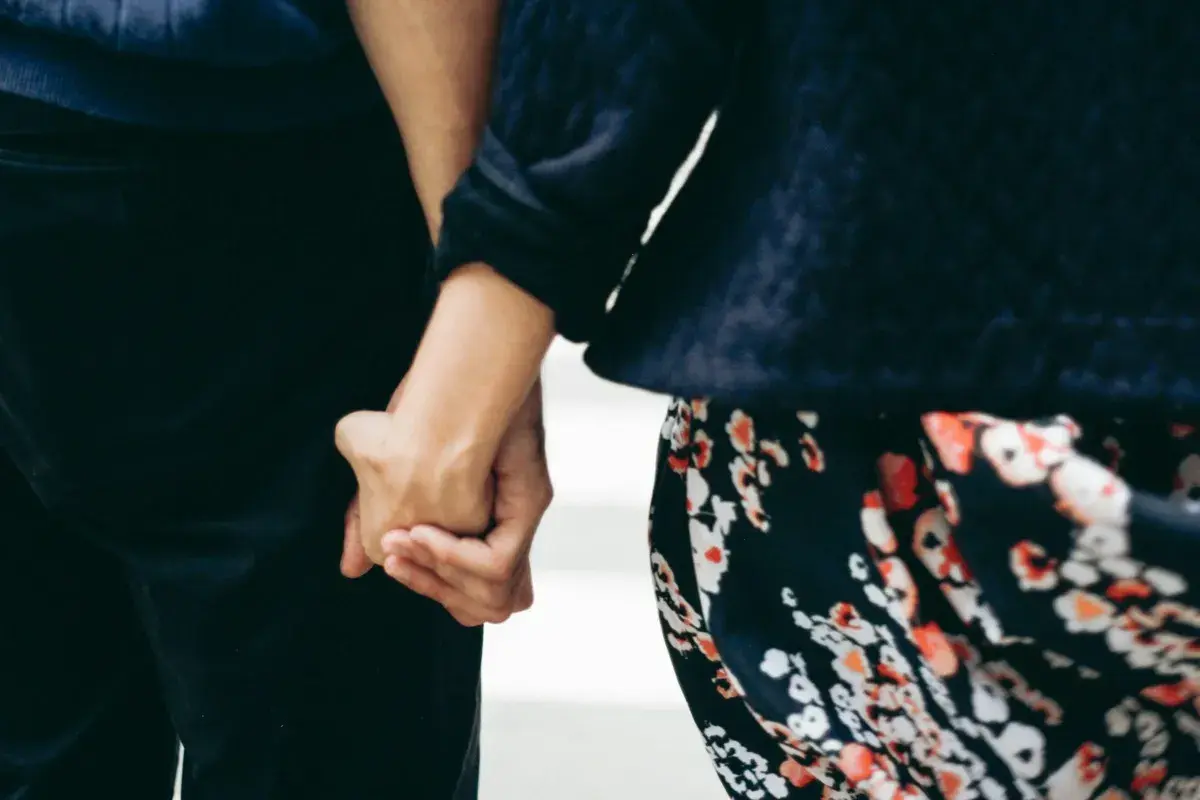Mani intrecciate, un gesto che parla di affinità di coppia e complicità. La donna indossa un abito floreale, l'uomo pantaloni scuri.