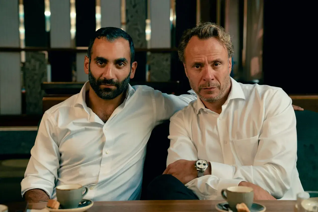 Två män i vita skjortor sitter vid ett bord med kaffe. En arm vilar på den andres axel. Scen från hundarna serie.