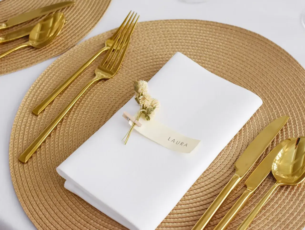 Elegante Tischdeko: Weiß gefaltete Servietten mit Namen "Laura", goldenes Besteck und kleine Trockenblumen auf einem geflochtenen Platzset.