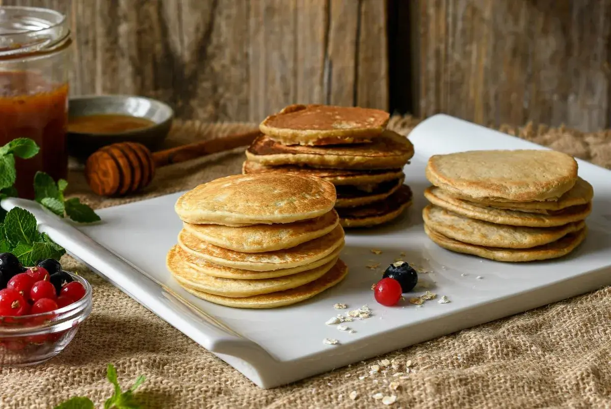 Montón de tortitas de avena doradas, servidas con bayas frescas, miel y menta.
