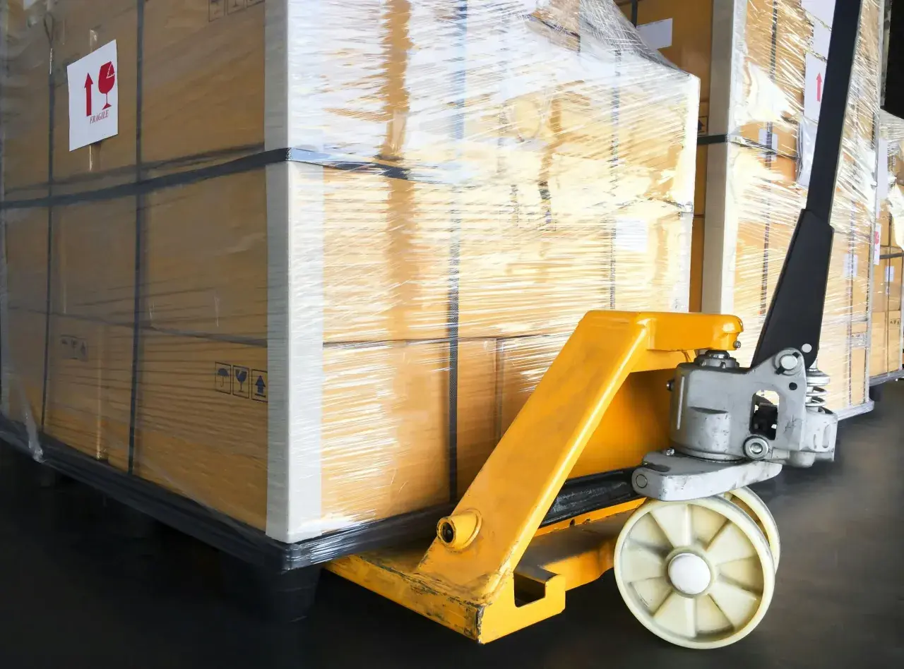 Wózek paletowy przygotowuje transport materaca. Duże, owinięte folią pudła czekają na załadunek.