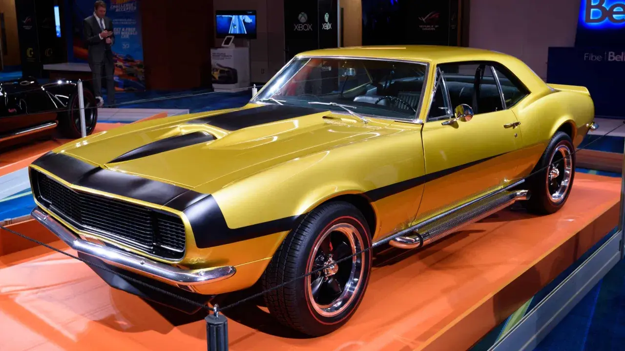 En glänsande gul Chevrolet Camaro SS 1969 med svarta ränder på huven och sidan. Bilen står på en orange yta.