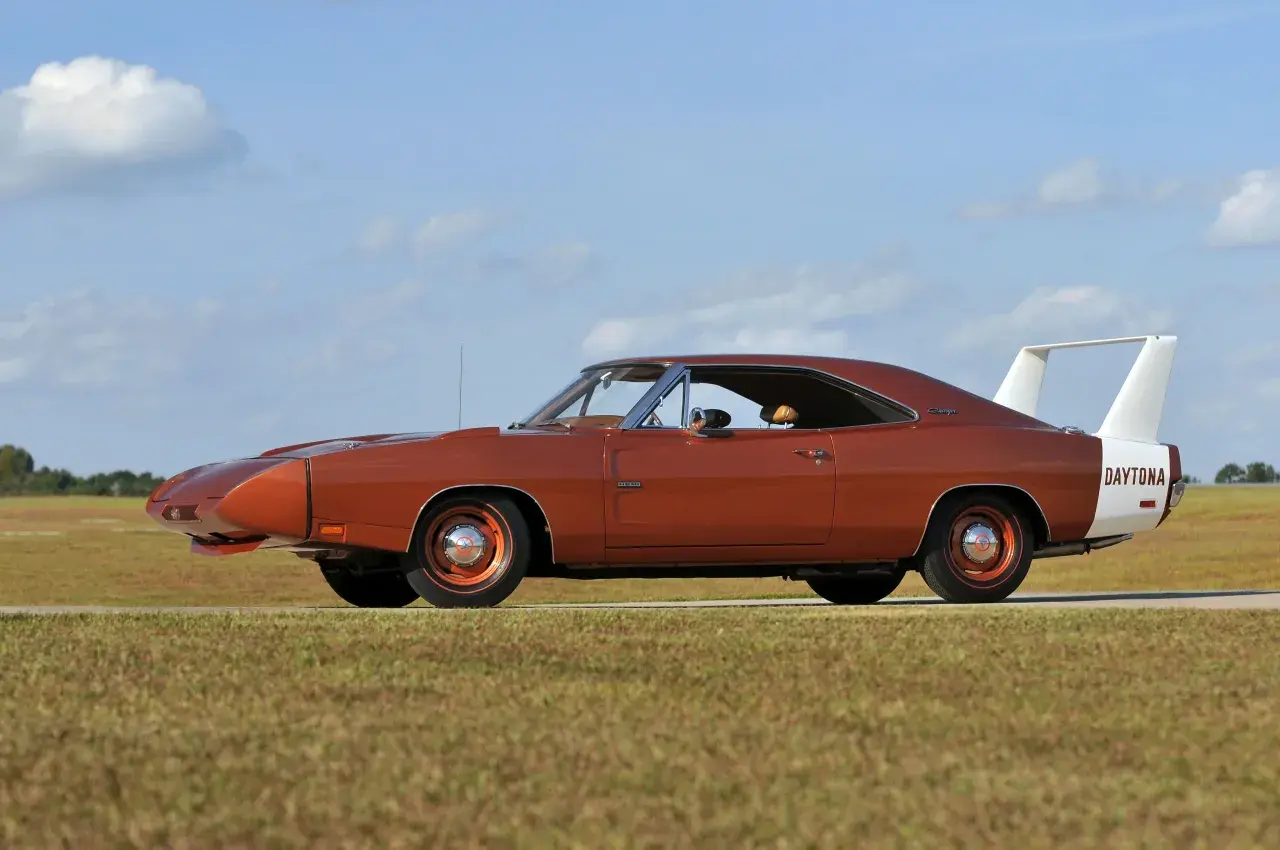 Pomarańczowy Dodge Daytona z charakterystycznym spojlerem i białym pasem z napisem "DAYTONA" na błotniku, zaparkowany na tle nieba z chmurami.