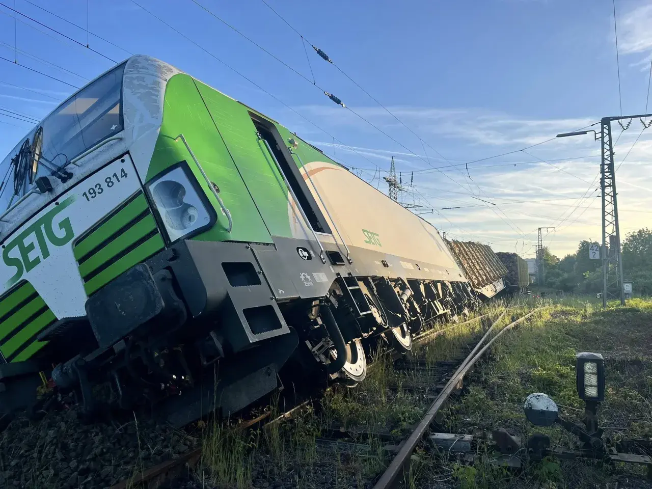 Zielony pociąg towarowy wykoleił się, przechylając się na bok na torach. W tle błękitne niebo i linie energetyczne.