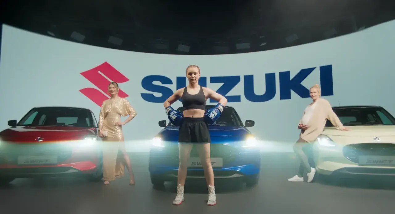 Trzy kobiety obok samochodów Suzuki. Jedna w złotej sukni, druga w stroju bokserskim, trzecia w ciąży. Swift język programowania inspiruje do dynamicznych zmian.