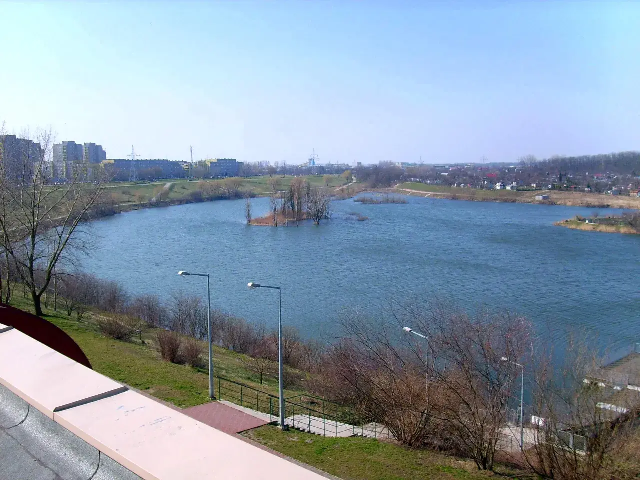 Spokojne jezioro Zatorze otoczone zielenią i zabudowaniami miejskimi. Na wodzie mała wyspa z drzewami.