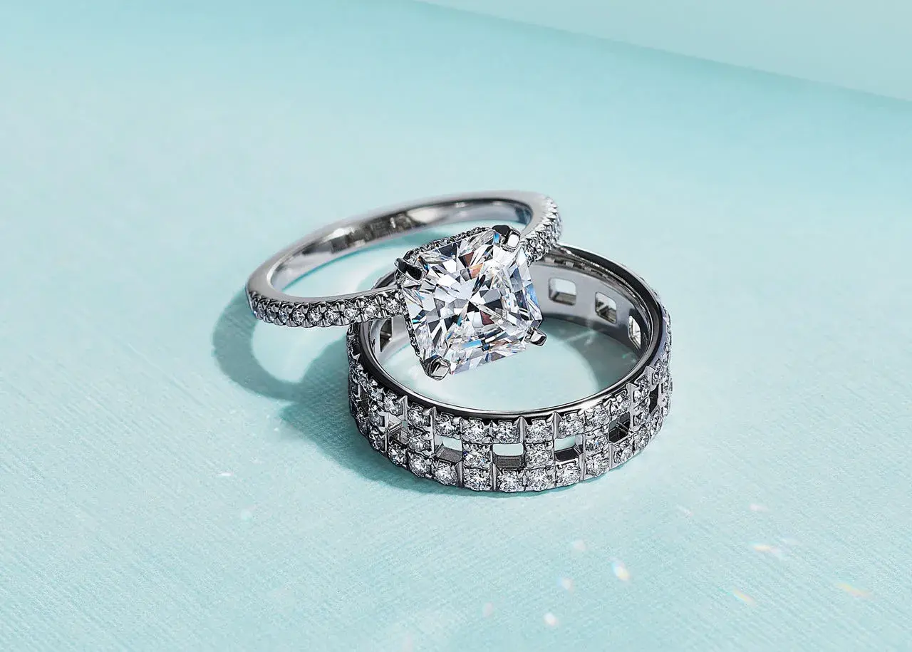 Un anillo de compromiso con un gran diamante cuadrado reposa sobre un anillo de matrimonio con incrustaciones de diamantes, ambos de plata.