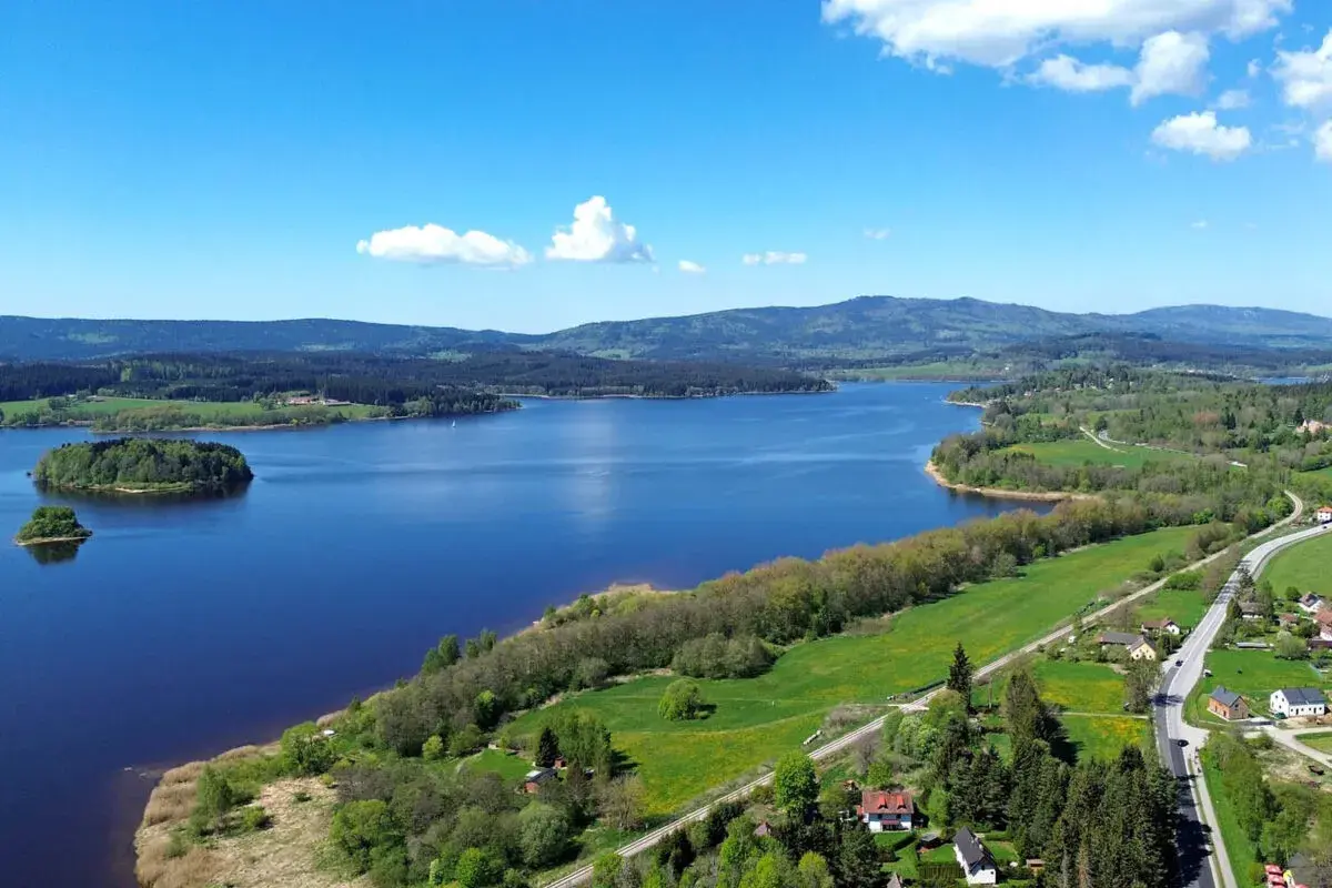Weite Sicht auf den Lipno Stausee mit bewaldeten Hügeln, grünen Wiesen und kleinen Häusern am Ufer.