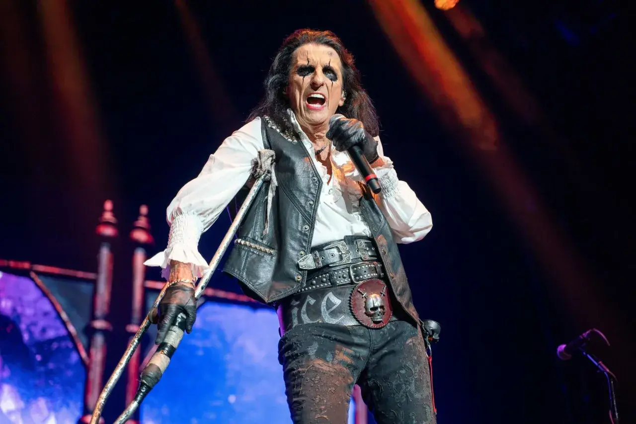 Alice Cooper na scenie z mikrofonem i kulami, w charakterystycznym makijażu i stroju.
