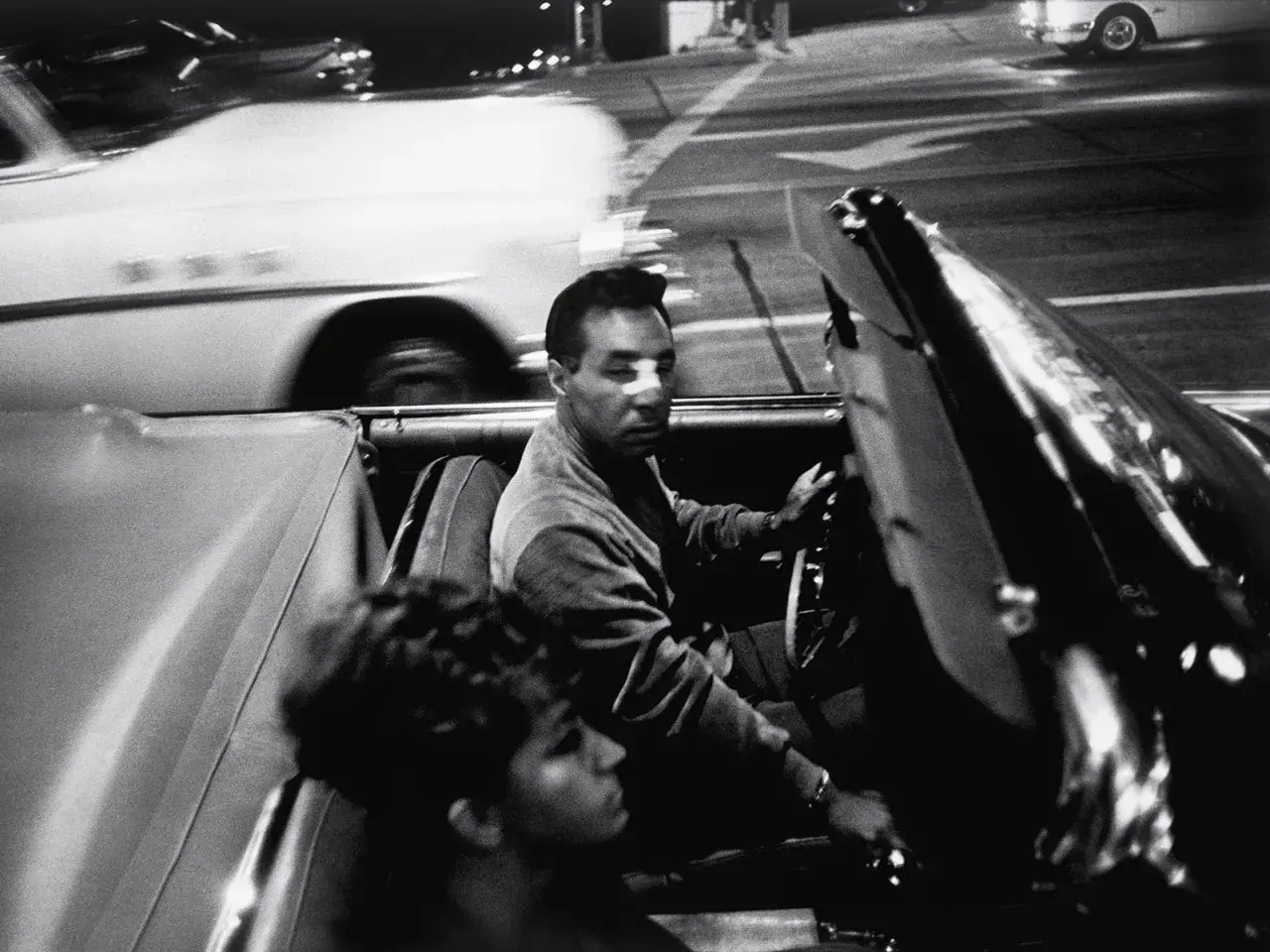 Garry Winogrand uchwycił nocną jazdę kabrioletem. Mężczyzna za kierownicą, kobieta obok. Rozmyte światła miasta.