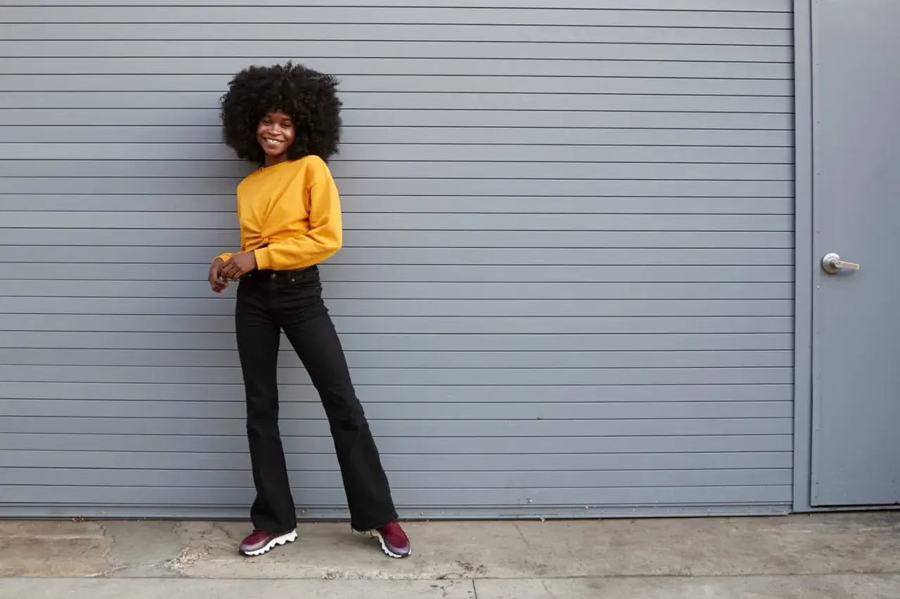 Une jeune femme souriante pose devant un mur gris, portant un haut jaune et un jean flare. Quelles chaussures avec jean flare ? Des baskets bordeaux complètent ce look décontracté.