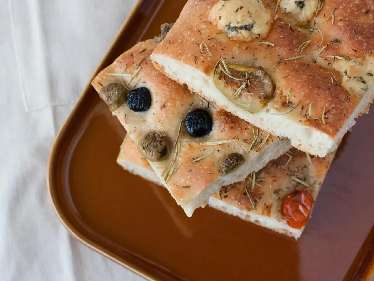 Focaccia Pomodoro mit Oliven und Rosmarin auf brauner Platte.