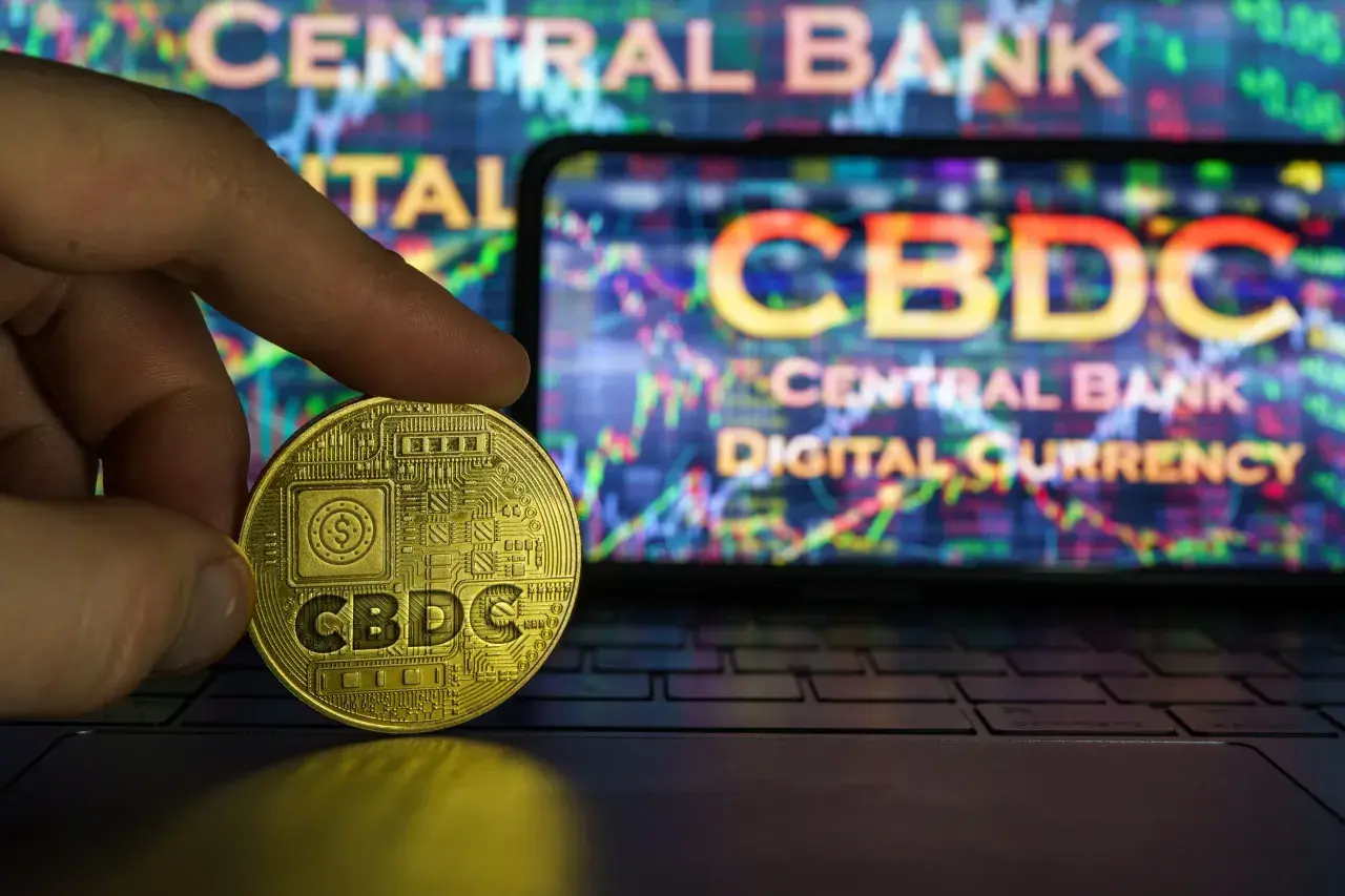 Złota moneta z napisem CBDC w dłoni na tle ekranu z wykresem i napisem "Central Bank Digital Currency". To przyszłość finansów, ale cbdc co to?