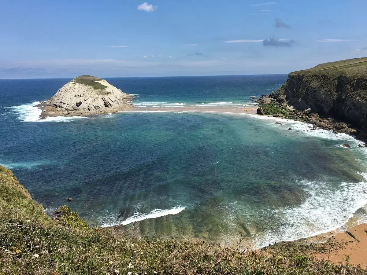 Vista de la Virgen del Mar en Santander, con una isla rocosa y aguas turquesas.