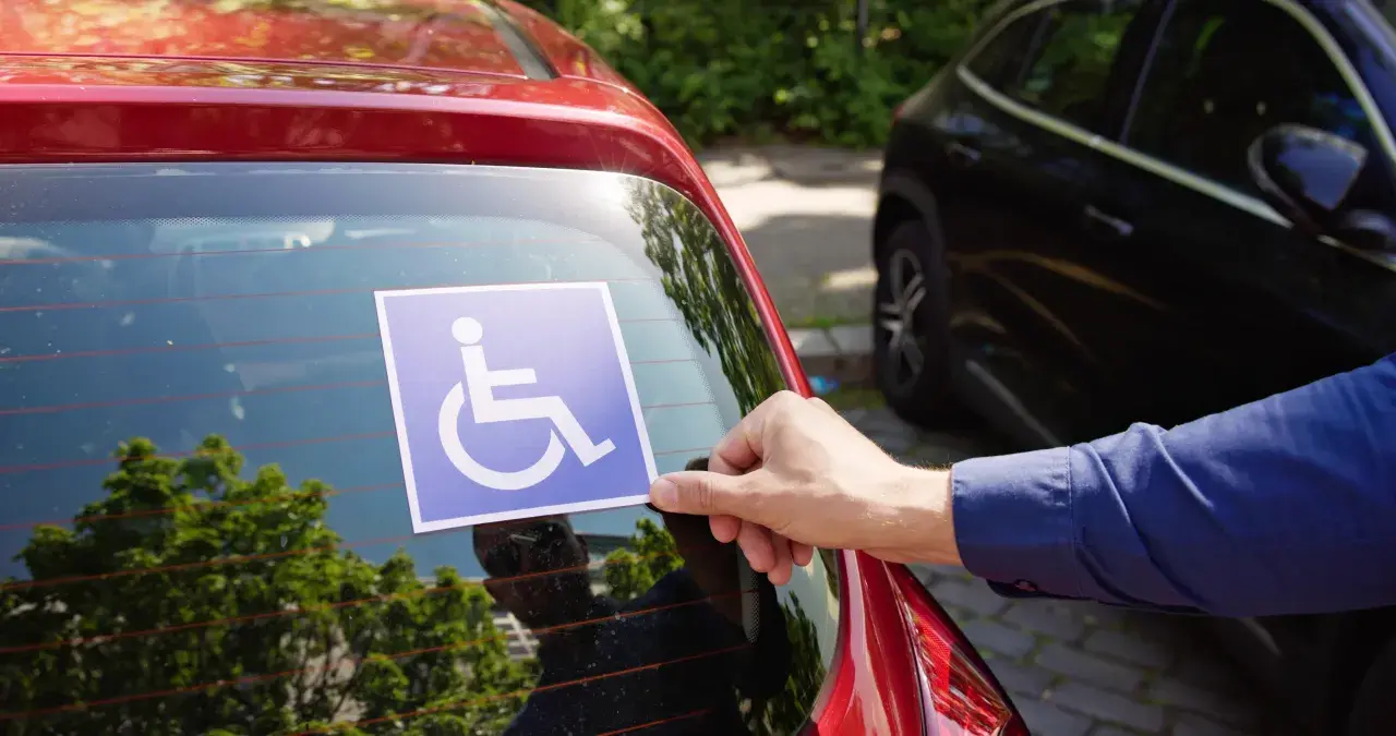 Ręka przykleja niebieską naklejkę z symbolem osoby na wózku inwalidzkim na tylnej szybie czerwonego samochodu. To przypomnienie o tym, jakie dokumenty do karty parkingowej są potrzebne.