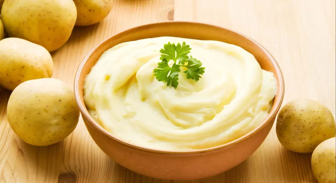Un tazón de cremoso puré de patatas, decorado con perejil, junto a patatas crudas. ¡Así es como hacer puré de patatas perfecto!