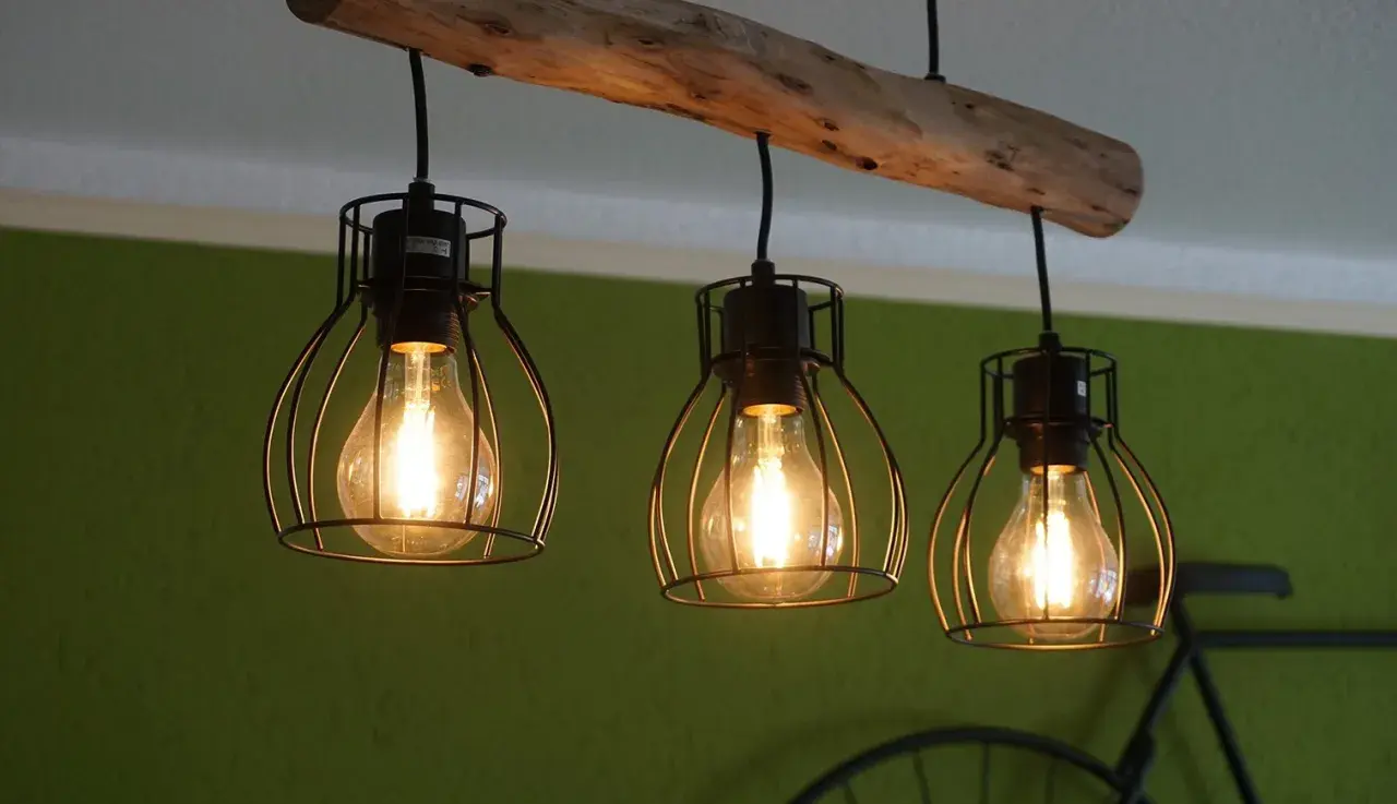 Lámpara de techo con tres bombillas Edison dentro de jaulas metálicas, colgadas de una rama de madera rústica.