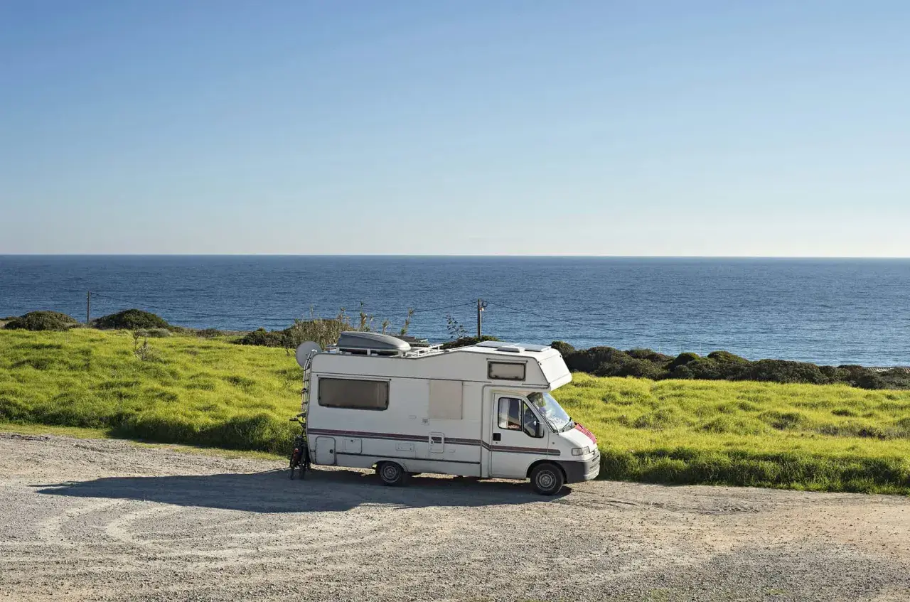 Camping-car blanc garé sur une aire gratuite en bord de mer en Bretagne. L'océan bleu s'étend à l'horizon, avec des champs verdoyants.