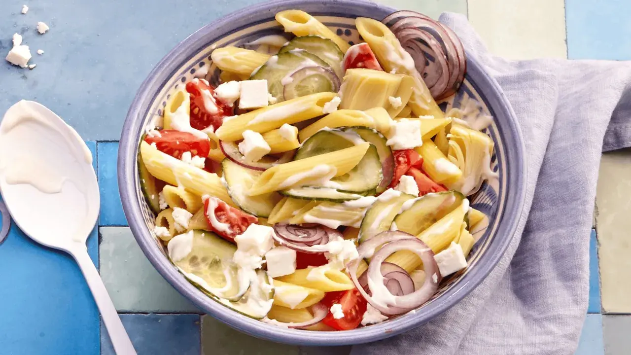 Schüssel mit griechischem Nudelsalat: Penne, Gurken, Tomaten, rote Zwiebeln und Feta in cremiger Soße.