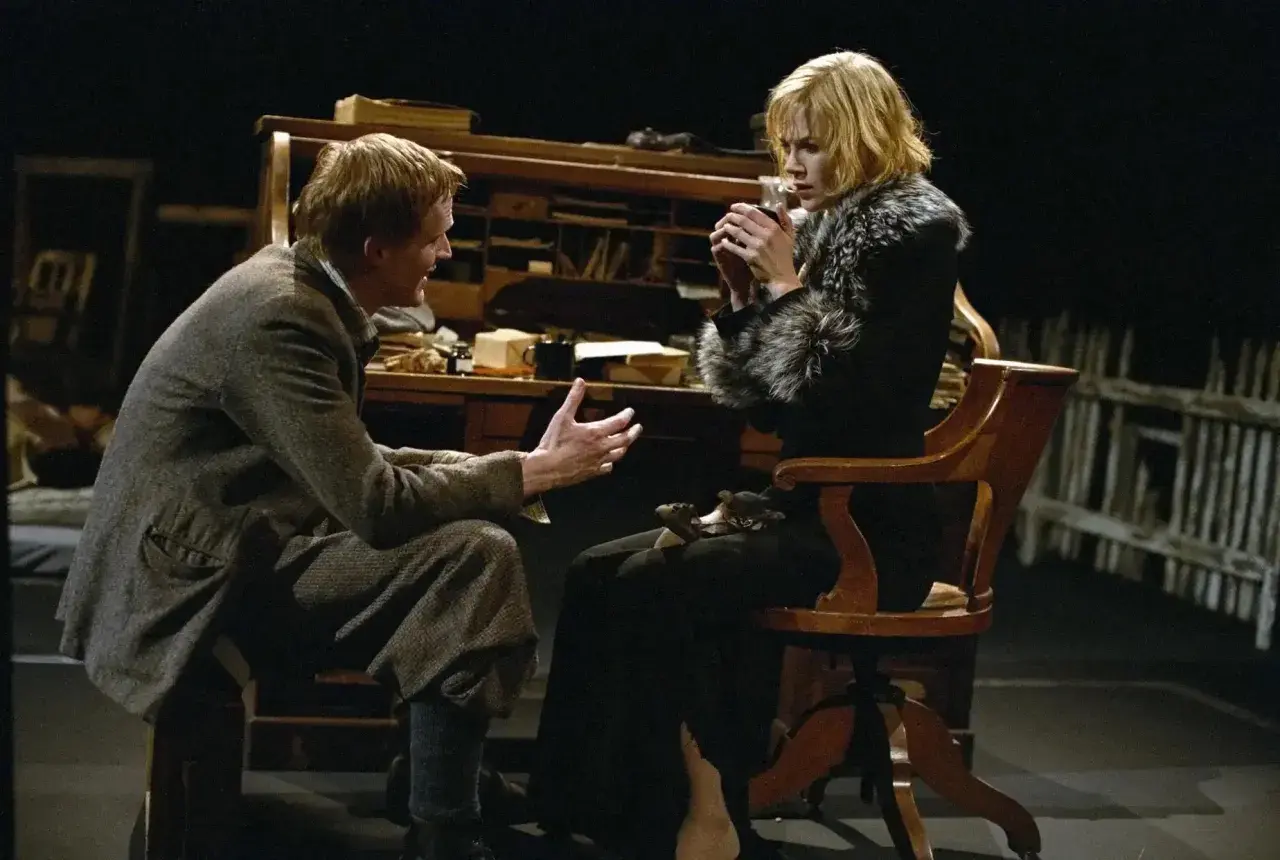 Una scena intensa da Dogville recensione: un uomo parla animatamente a una donna seduta, che tiene una tazza tra le mani.