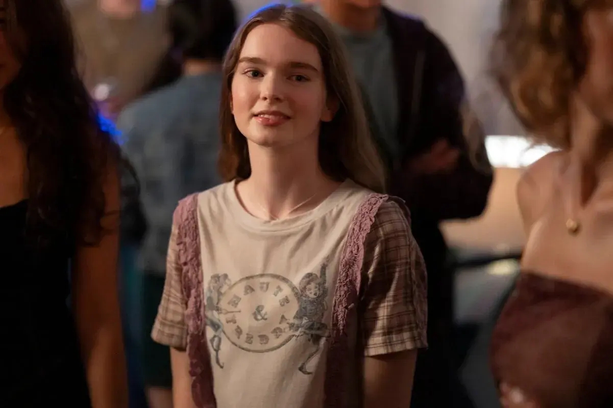 Młoda dziewczyna w koszulce z nadrukiem, przypominająca postać z filmów Chloë Grace Moretz, uśmiecha się na imprezie.