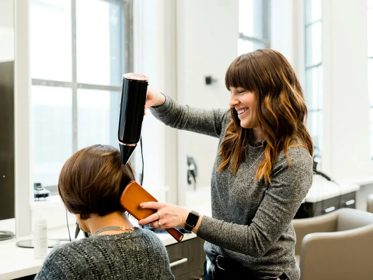 Estilista sonríe mientras seca el cabello de una clienta. Un buen momento para cortarse el pelo es cuando te sientes renovada.