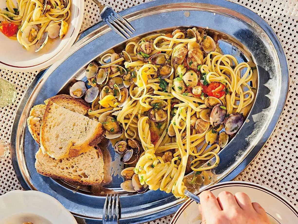 En stor silverbricka med pasta vongole, musslor, körsbärstomater och vitlöksbröd. En hand tar en gaffel med pasta.