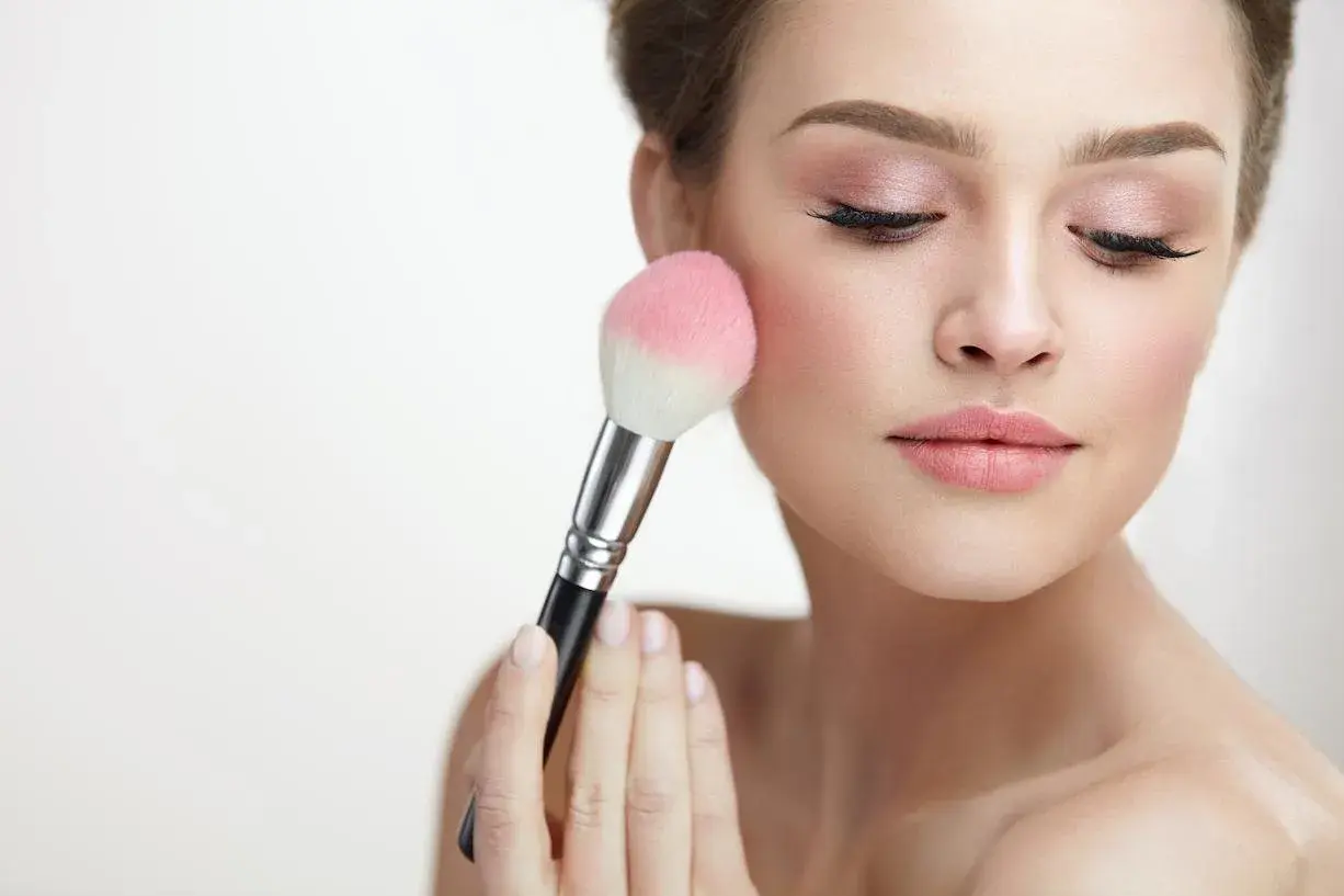 Mujer aplicando rubor en sus mejillas con una brocha. El tono rosado del rubor realza su piel, creando un look natural y radiante.