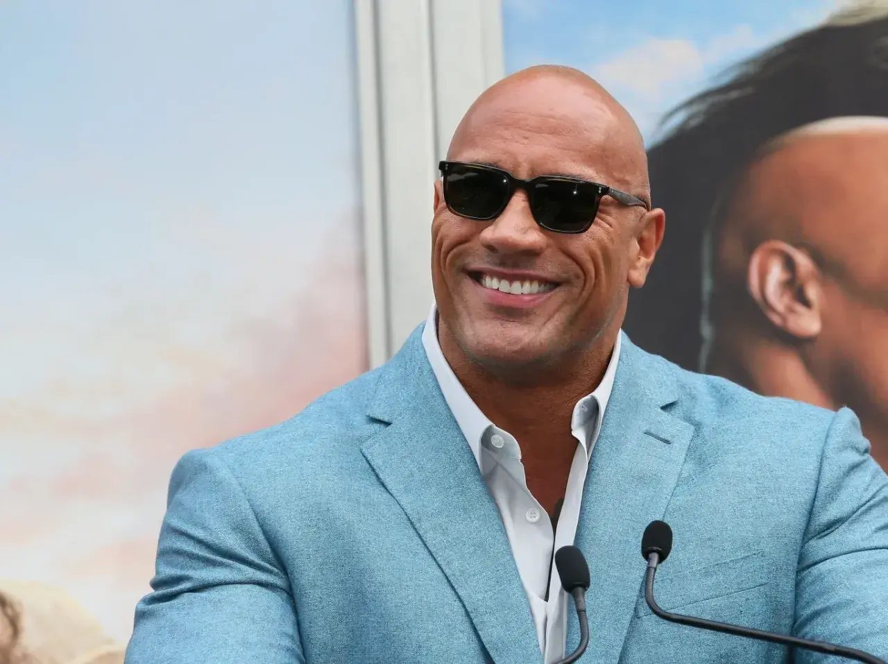 Dwayne "The Rock" Johnson, być może najbogatszy aktor na świecie, w błękitnej marynarce i okularach przeciwsłonecznych, uśmiecha się do kamery.