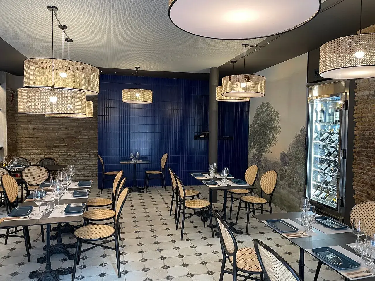 Interior de la brasserie 1885, con mesas puestas, sillas de mimbre y lámparas de ratán. Pared azul y mural de paisaje.