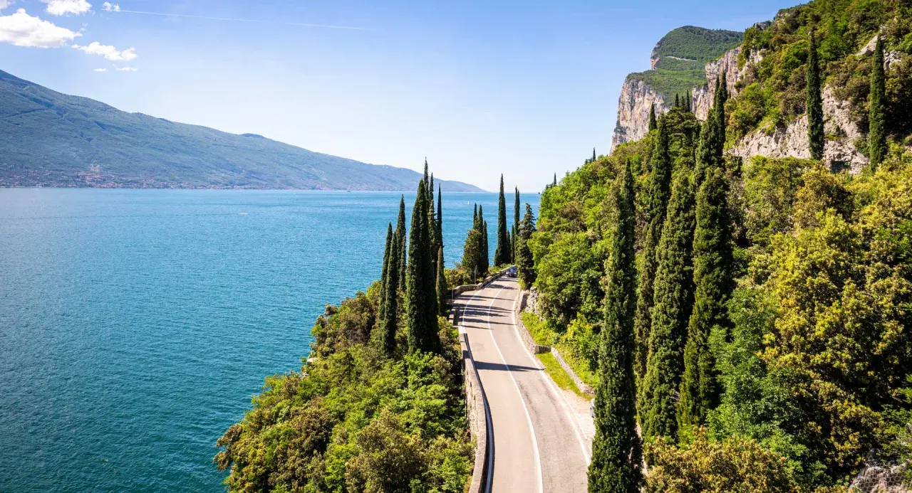Route panoramique longeant un lac italien, idéale pour le camping-car. Cyprès élancés, falaises et eau bleue invitent à l'aventure.