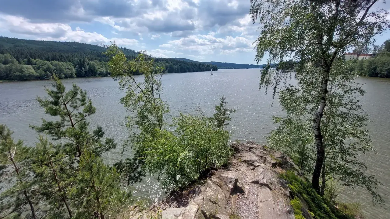 Felsiger Uferrand am Moldau Stausee mit Bäumen und Segelbooten auf dem glitzernden Wasser unter bewölktem Himmel.
