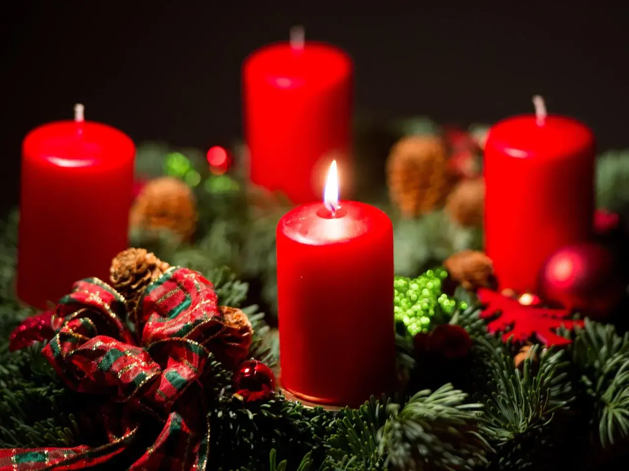 Ein Adventskranz mit vier roten Kerzen, Tannenzweigen, Zapfen und Schleifen. Die erste Kerze brennt und symbolisiert, warum man Advent feiert: die Vorfreude auf Weihnachten.